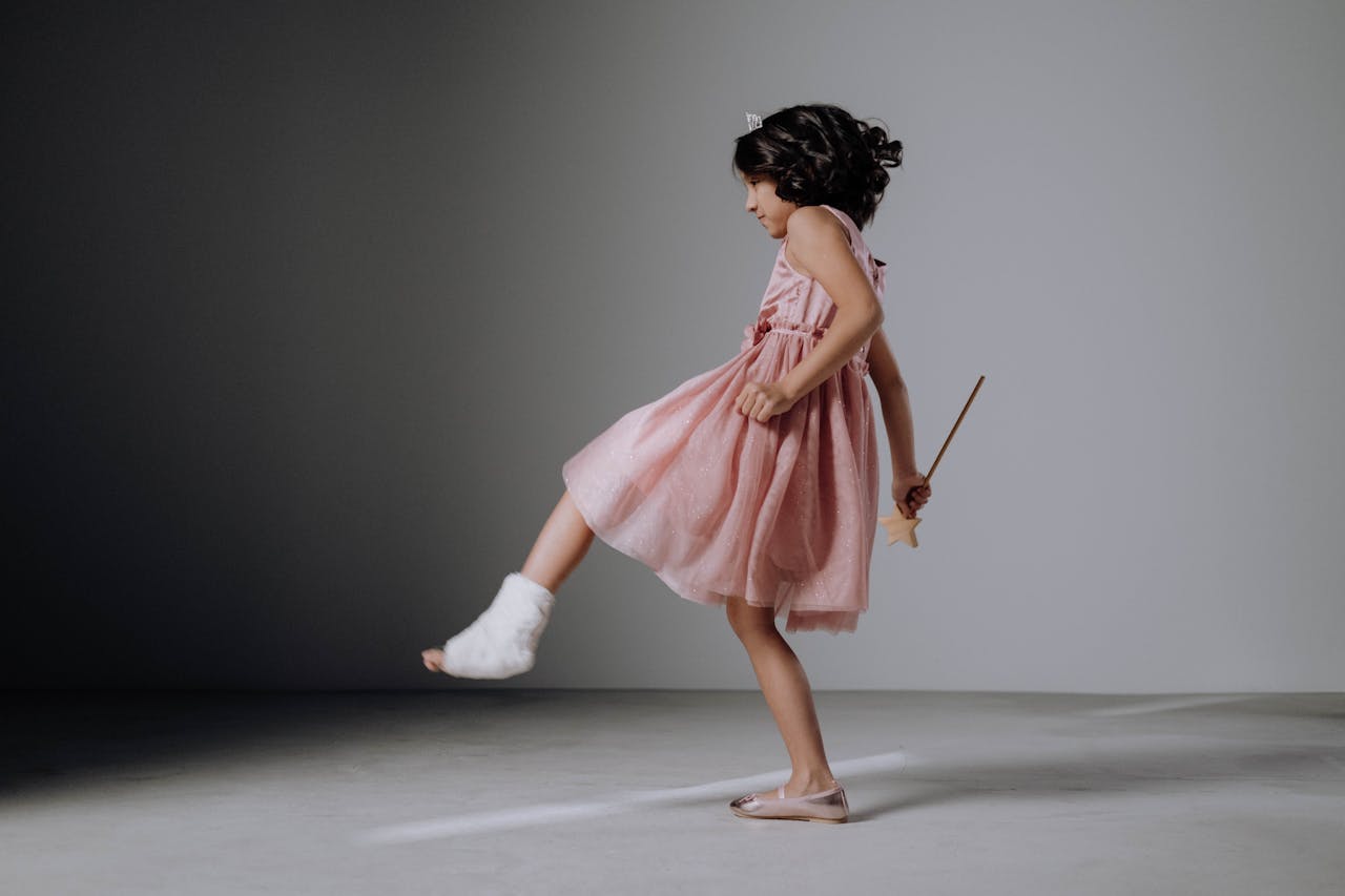Child in pink dress and orthopedic cast kicking playfully in a studio setting.