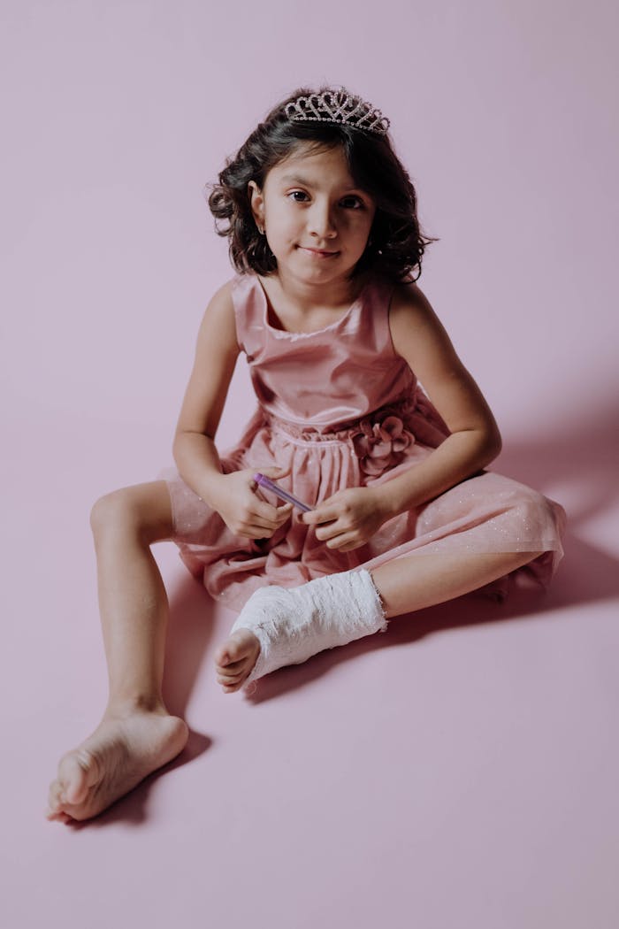 A young girl in a pink dress and tiara smiles while sitting with a leg cast in a studio.
